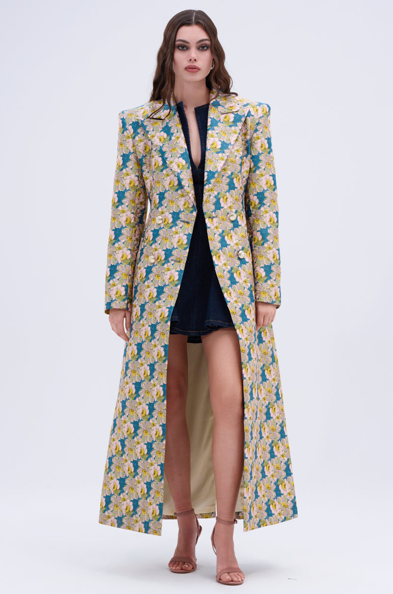 A woman with wavy hair faces forward in the SCARLETT LUXE TRENCH IN BROCADE, styled over a dark mini dress and strappy nude heels, set against a simple, light background.
