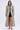 A woman with wavy hair faces forward in the SCARLETT LUXE TRENCH IN BROCADE, styled over a dark mini dress and strappy nude heels, set against a simple, light background.