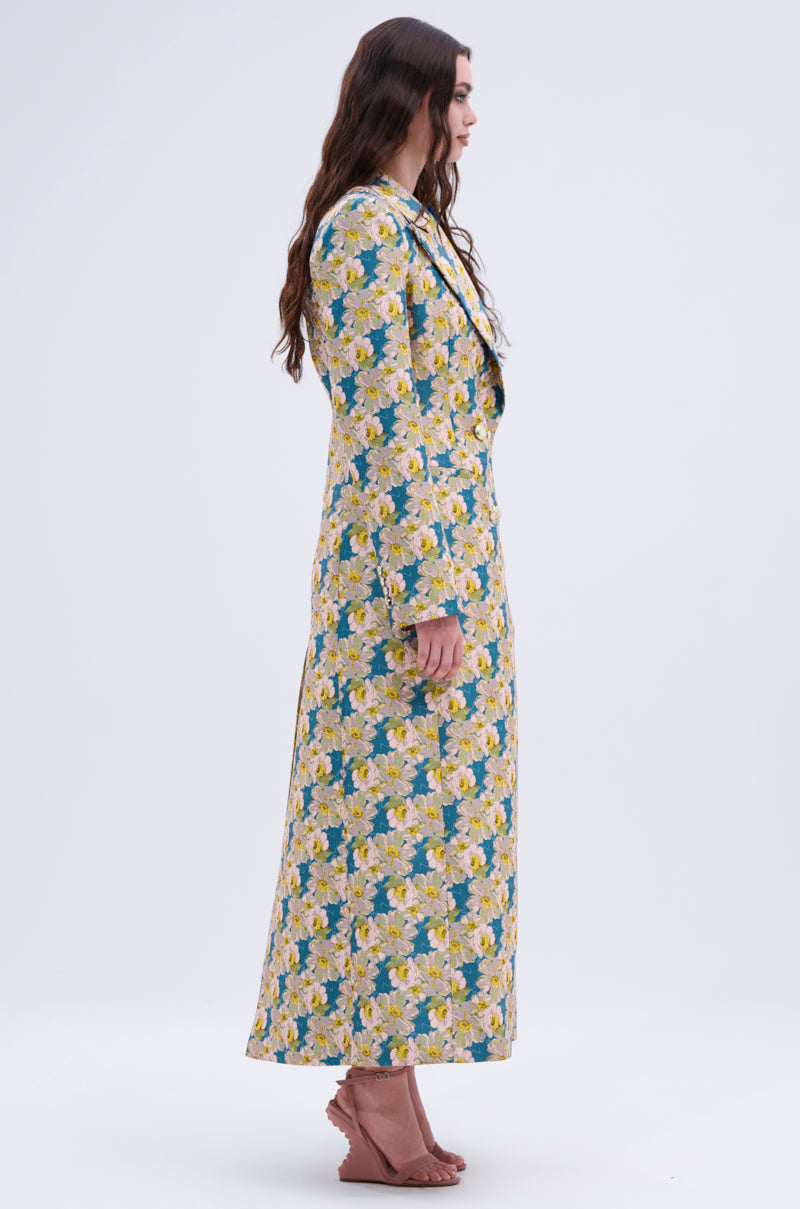A woman in profile wears the SCARLETT LUXE TRENCH IN BROCADE, a long, floral-patterned coat with yellow and white flowers and green leaves. She has wavy brown hair and light brown sandals against a plain background.