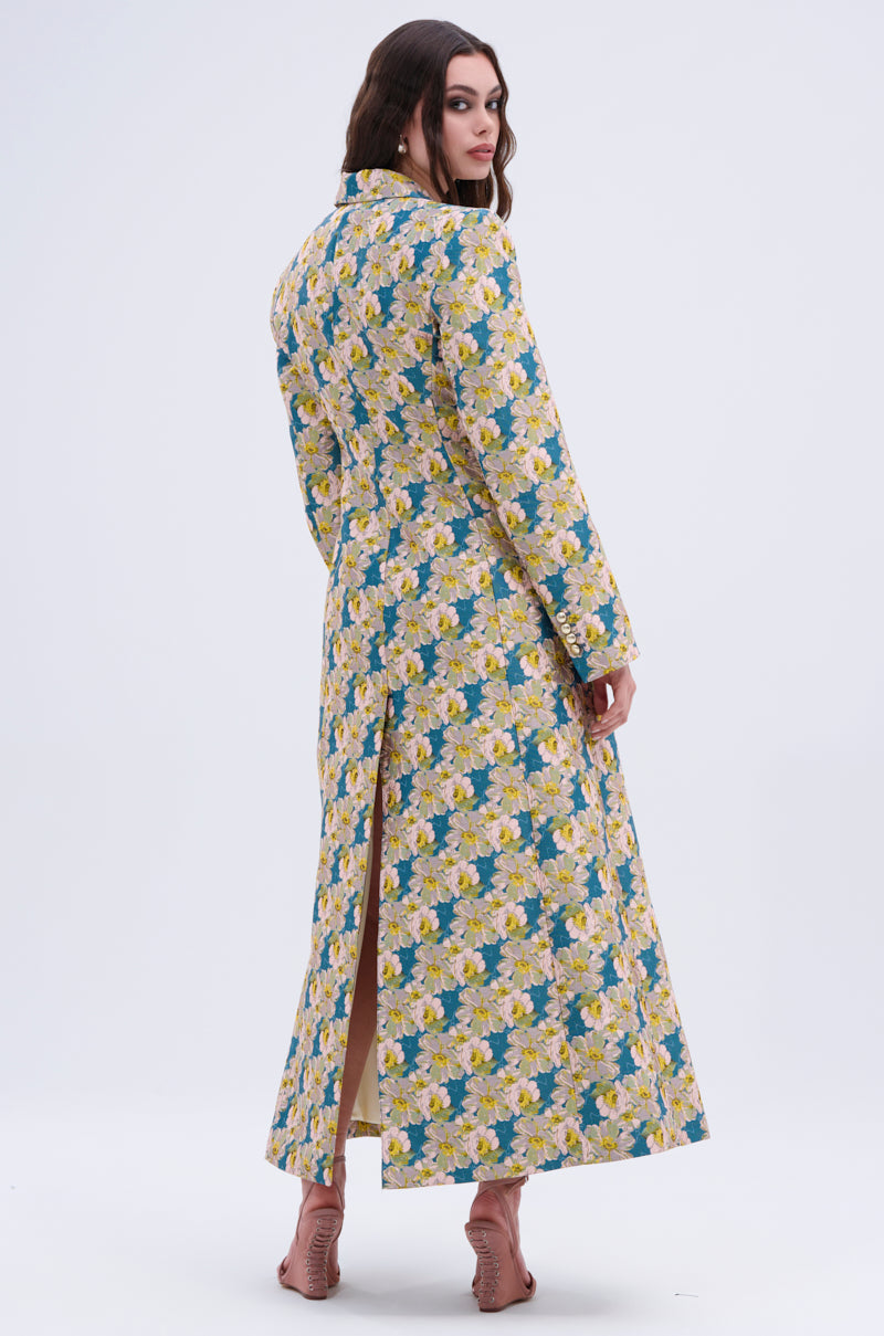 A woman poses with her back to the camera in the SCARLETT LUXE TRENCH IN BROCADE, which features yellow and white floral brocade, a back slit, and is styled with pink open-toed heels against a plain backdrop.