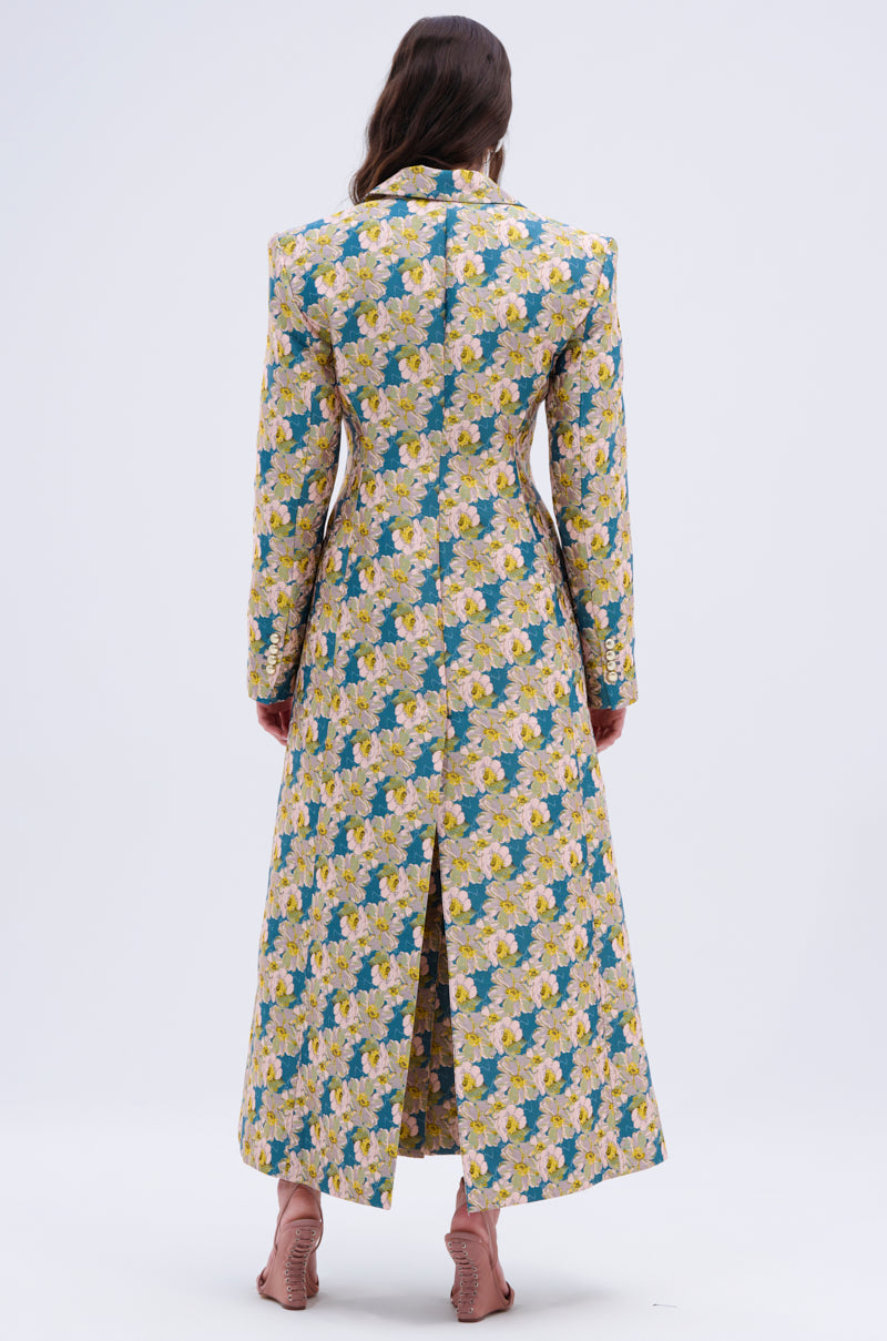 A woman faces away wearing the SCARLETT LUXE TRENCH IN BROCADE—a long, fitted coat with yellow and pink floral brocade on blue, long sleeves, a back slit, and paired with pink heeled sandals.