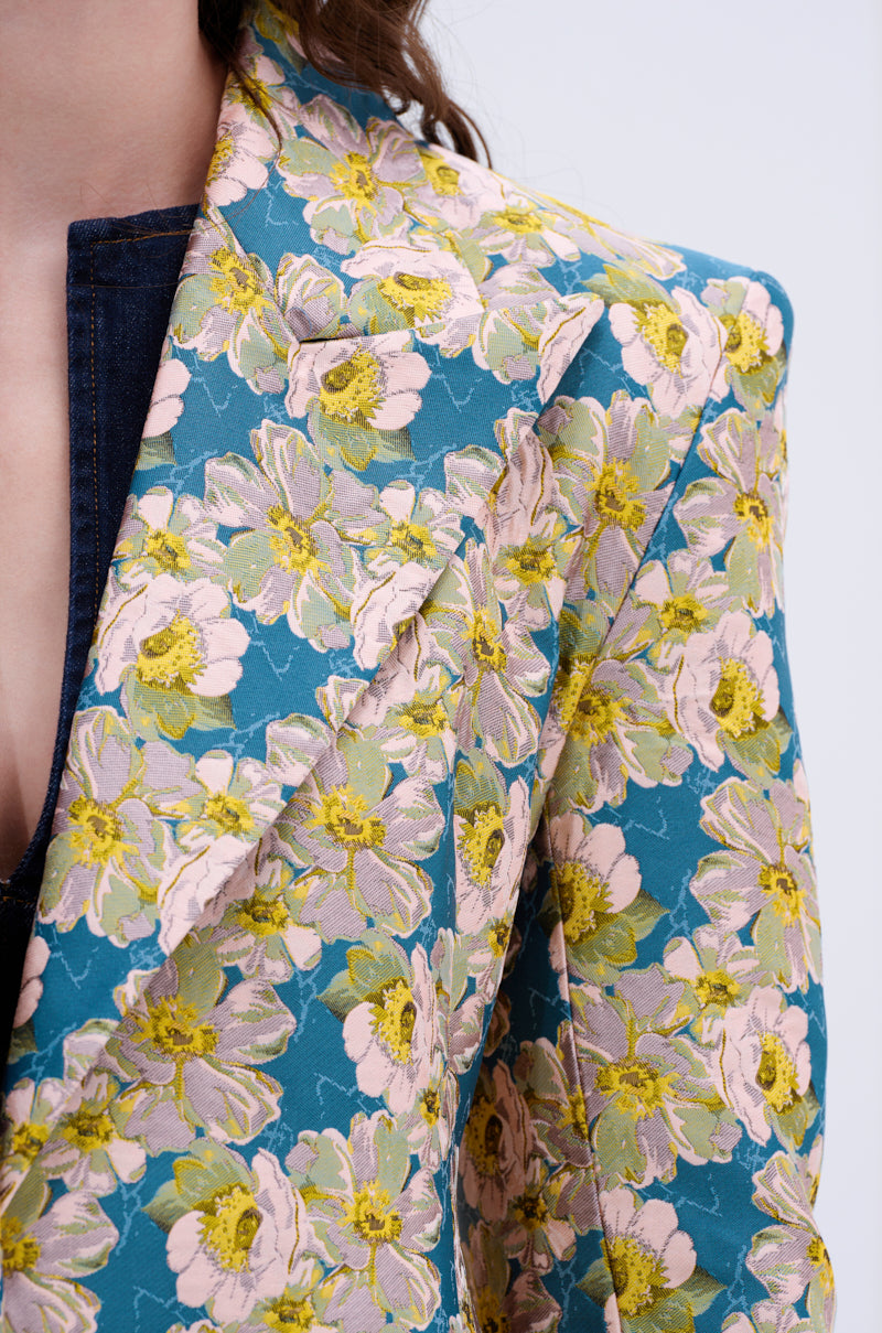 Close-up of a person wearing the SCARLETT LUXE TRENCH IN BROCADE, a teal blazer featuring bold floral brocade with large yellow, green, and pink flowers, layered over a dark blue or black denim inner garment. Partial face and body visible.