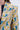 Close-up of a person wearing the SCARLETT LUXE TRENCH IN BROCADE, a teal blazer featuring bold floral brocade with large yellow, green, and pink flowers, layered over a dark blue or black denim inner garment. Partial face and body visible.