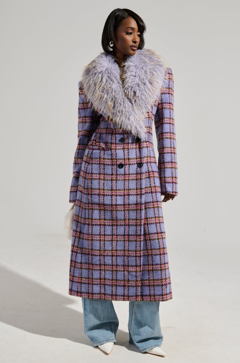 A person poses in the TEA FOR TWO FUR TRIM TRENCH, a long purple plaid coat featuring a satin interior lining and an oversized fur collar. They pair it with wide-legged jeans and white pointed shoes against a plain, light gray background.