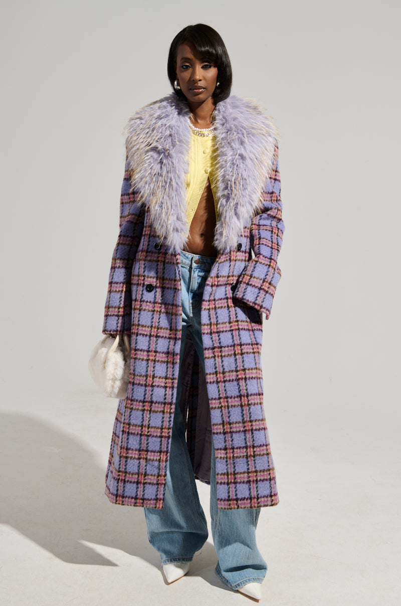 Wearing the TEA FOR TWO FUR TRIM TRENCH, a stylish plaid coat with a luxurious fur trim, this person pairs it with a yellow shirt, wide-leg jeans, and white pointed-toe shoes. Holding a fluffy white handbag, they stand against a plain background, exuding chic elegance.