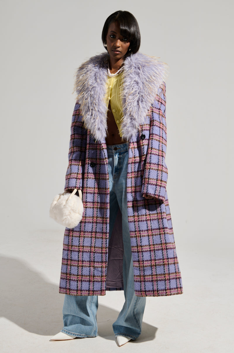 A person dons the TEA FOR TWO FUR TRIM TRENCH, a long plaid coat with a satin interior lining and a large faux fur collar. They pair it with a yellow top and blue jeans, accessorizing with a small white handbag and white pointed shoes, standing against a plain background.