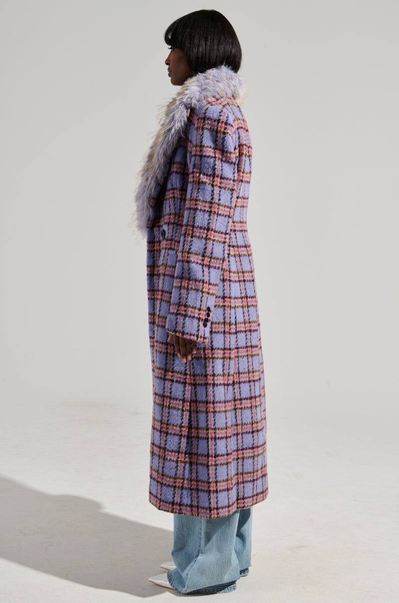 A person stands in profile wearing the TEA FOR TWO FUR TRIM TRENCH, a long coat with a purple plaid design and satin interior lining. They have straight, dark hair and are dressed in light blue jeans and white pointed shoes, set against a plain gray background.
