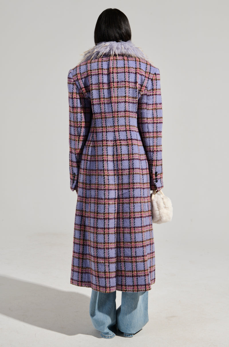 A person with long dark hair stands facing away, sporting the TEA FOR TWO FUR TRIM TRENCH in a purple plaid pattern with a luxurious satin interior lining. They accessorize with a small fluffy white handbag and blue jeans beneath, set against a plain light gray background.