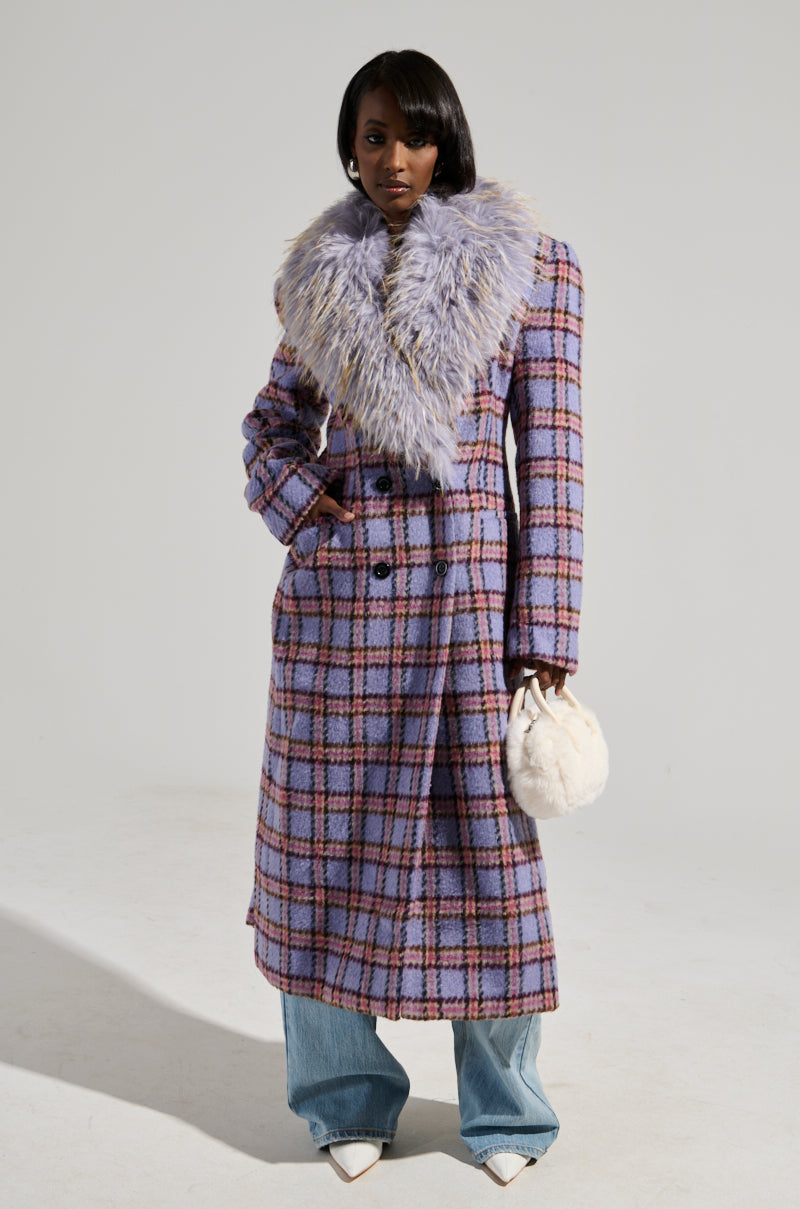A person models the TEA FOR TWO FUR TRIM TRENCH, featuring a purple plaid design with a luxurious, fluffy collar and satin interior lining. They are carrying a small white bag and wearing denim jeans along with white pointed shoes against a plain, light-colored backdrop.
