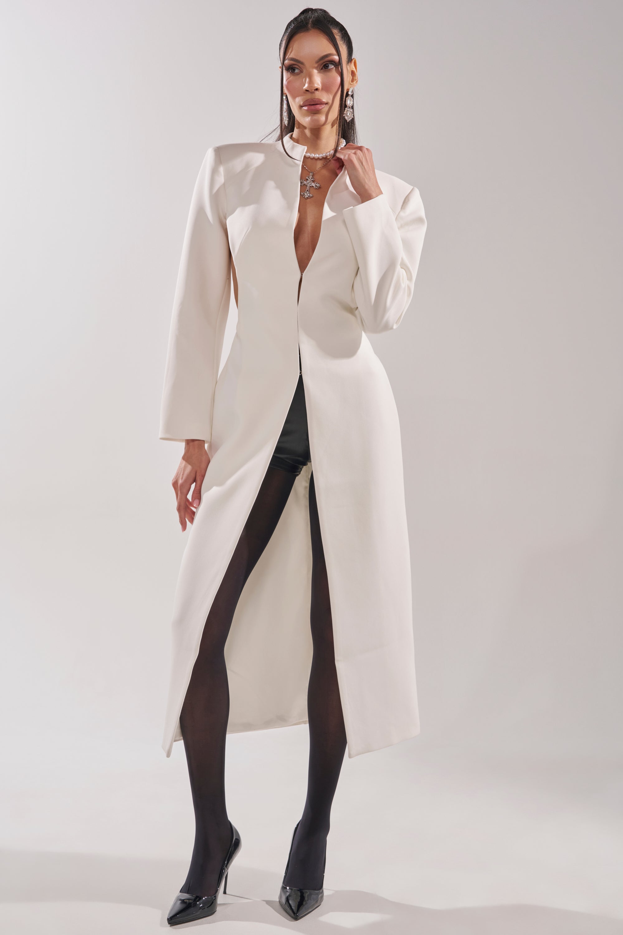 A woman poses confidently against a plain light background, wearing the WHEREVER YOU ARE CUTOUT TRENCH IN WHITE styled with black tights, pointed-toe heels, and a statement necklace. 0