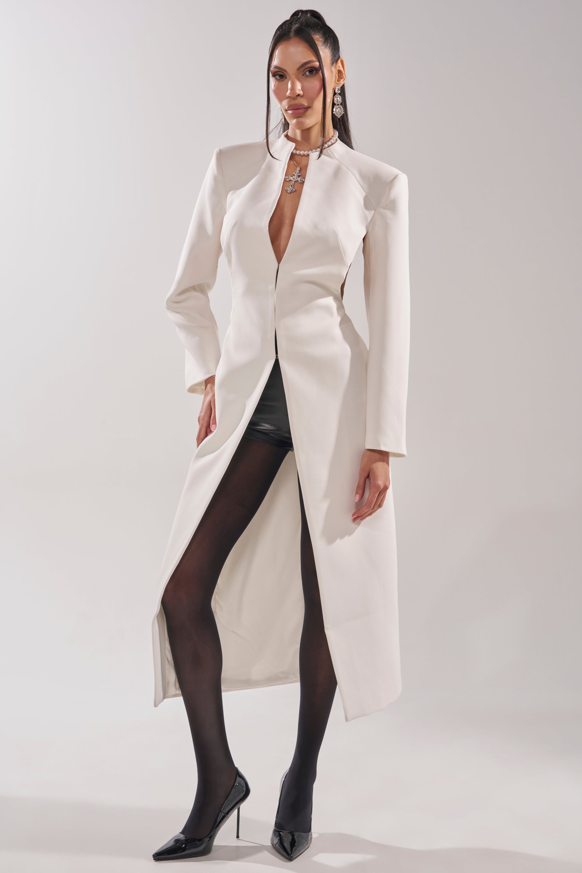 A woman poses against a light background wearing the WHEREVER YOU ARE CUTOUT TRENCH IN WHITE, styled with black shorts, sheer tights, black high heels, and a bold silver necklace. 1