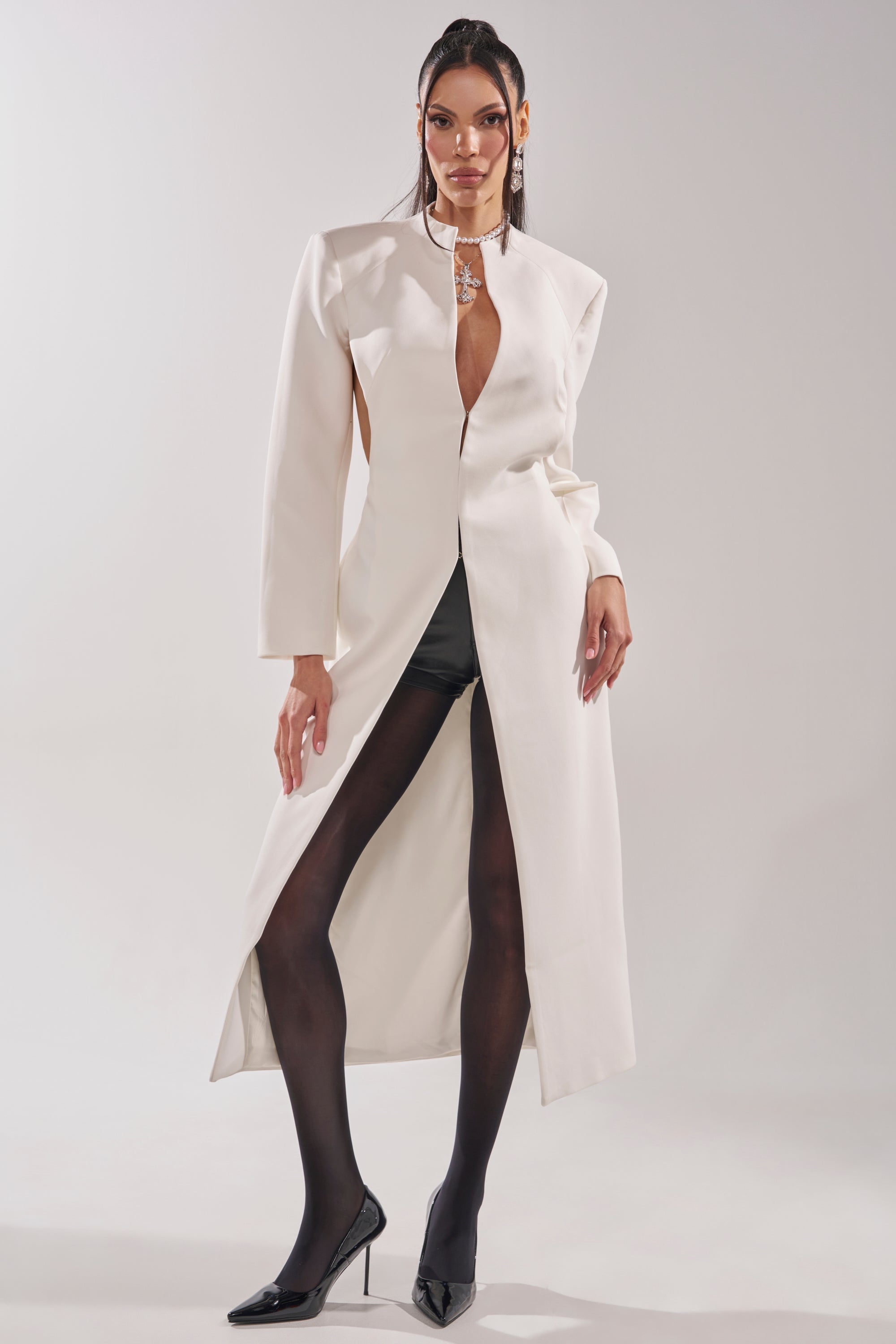 A model poses in the WHEREVER YOU ARE CUTOUT TRENCH IN WHITE, styled with black sheer tights and black pointed high heels against a plain light background. 3