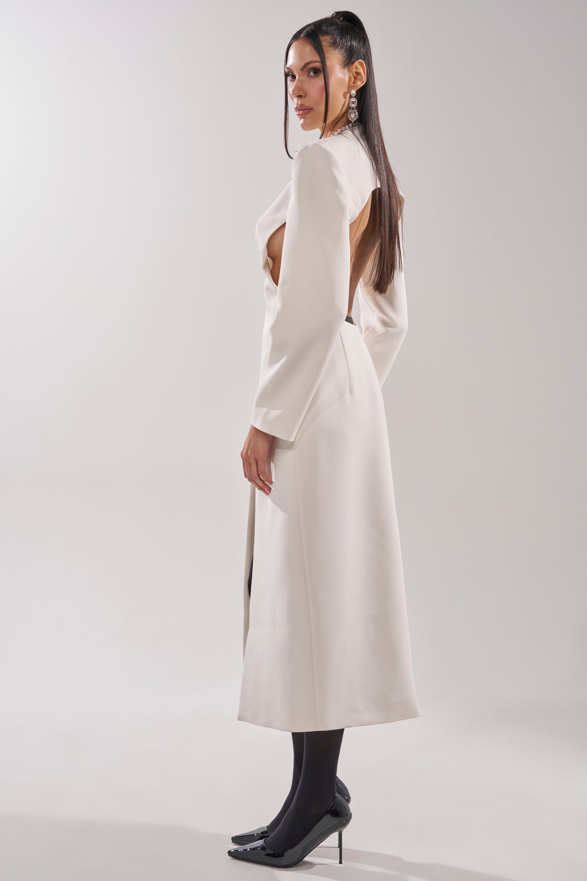A woman with long dark hair stands sideways against a plain light background, wearing the WHEREVER YOU ARE CUTOUT TRENCH IN WHITE, styled with black tights and black high heels. 4