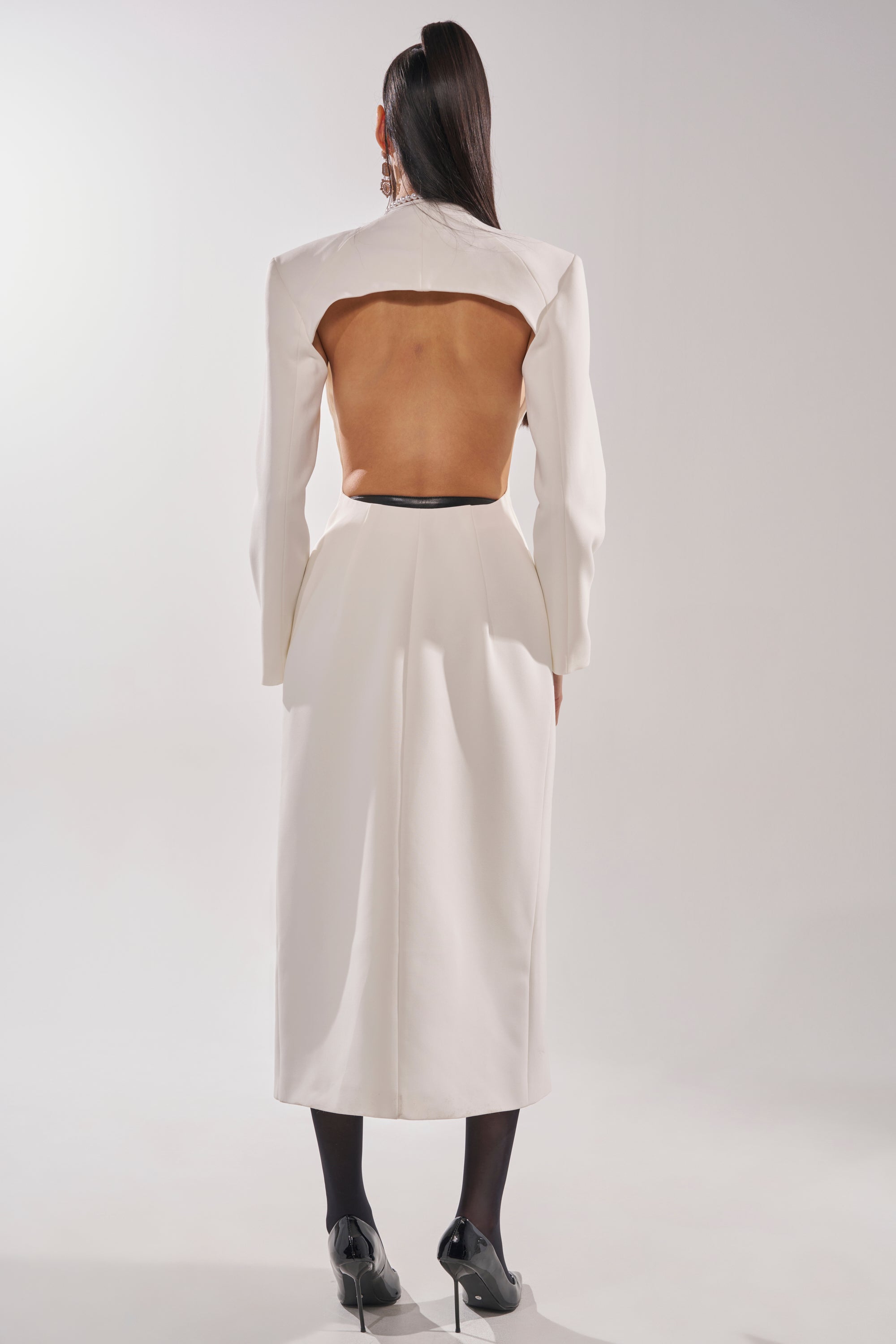 A person with long dark hair stands facing away against a plain light background, wearing the WHEREVER YOU ARE CUTOUT TRENCH IN WHITE styled as a dress, paired with black stockings and high heels. 5