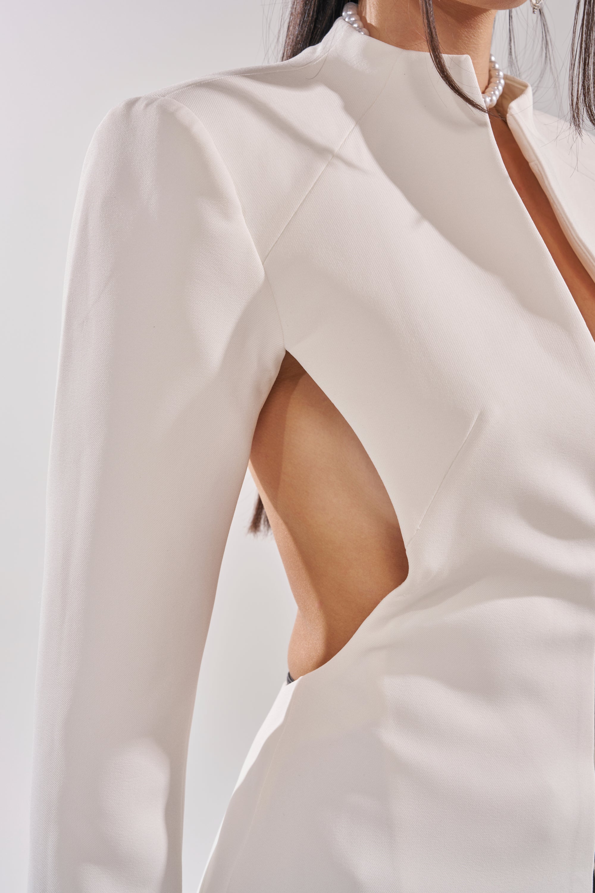 A woman models the WHEREVER YOU ARE CUTOUT TRENCH IN WHITE, featuring long sleeves and a side cutout revealing part of her torso. She has straight brown hair and accessorizes with a pearl necklace. 7