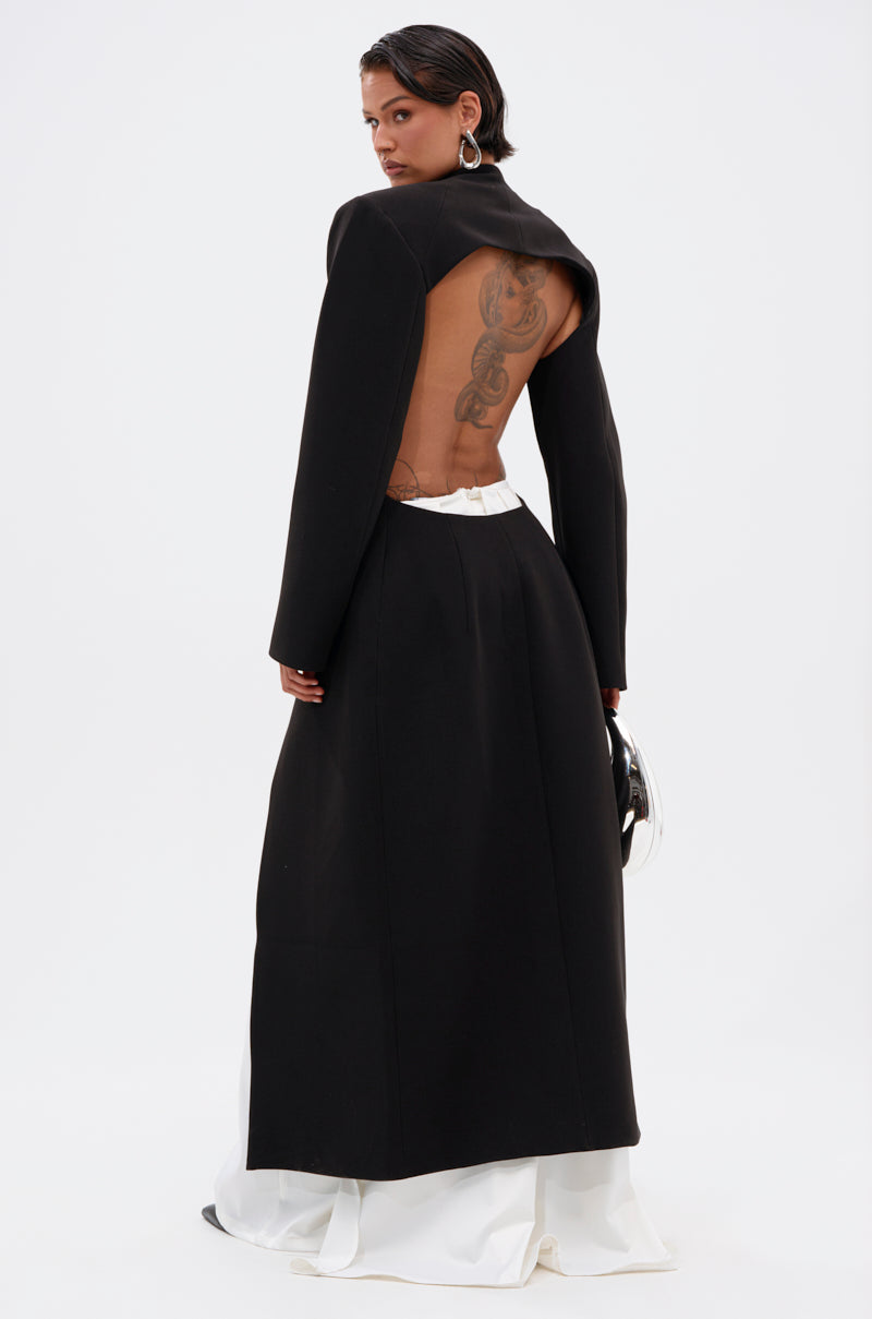 A woman faces away in the WHEREVER YOU ARE CUTOUT TRENCH, worn as a long black backless dress with long sleeves. Tattoos are visible, and a white skirt peeks out below. She holds a small bag in her left hand.