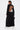 A woman faces away in the WHEREVER YOU ARE CUTOUT TRENCH, worn as a long black backless dress with long sleeves. Tattoos are visible, and a white skirt peeks out below. She holds a small bag in her left hand.