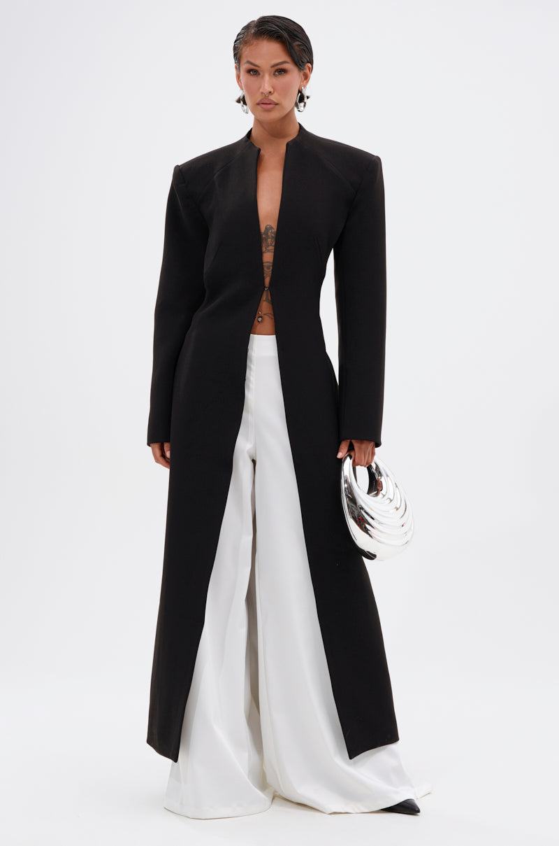 A person with short dark hair wears the WHEREVER YOU ARE CUTOUT TRENCH, styled open over wide white pants, and holds a shiny silver clutch. The modern, minimalist look stands out against a plain white background.