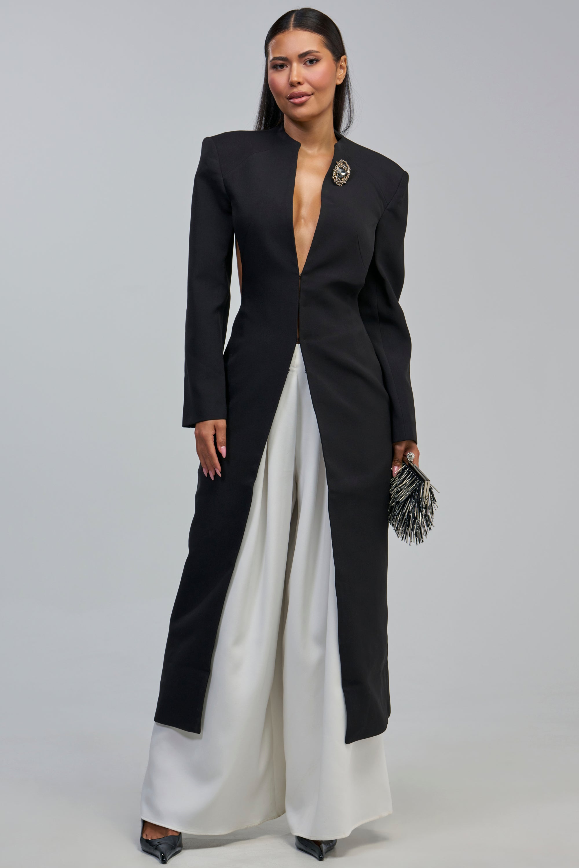 A woman wearing the WHEREVER YOU ARE CUTOUT TRENCH styled as an evening coat with a brooch, paired with white wide-leg pants and holding a black clutch with silver fringes, stands against a plain gray background.
