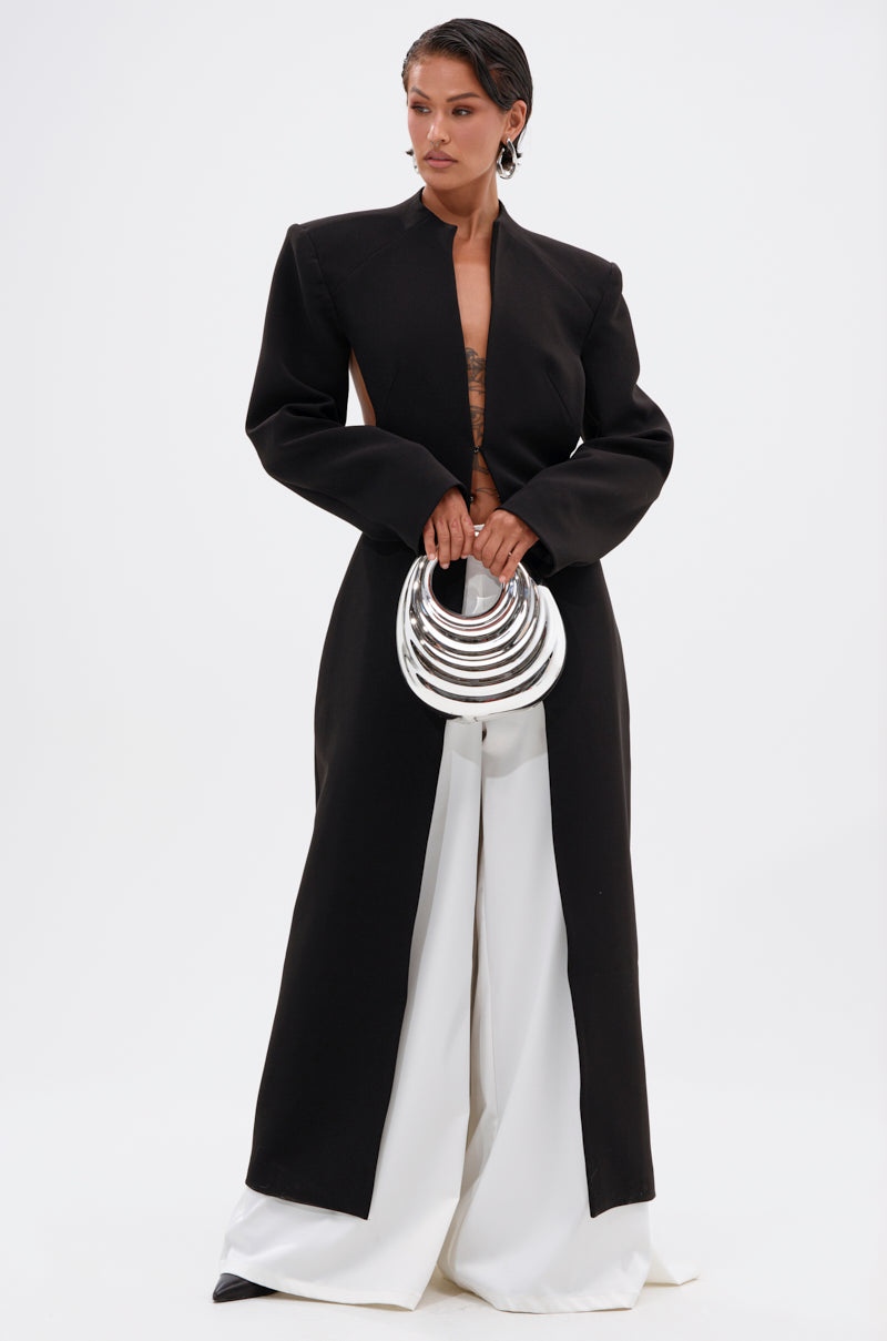 A person with short hair wears the WHEREVER YOU ARE CUTOUT TRENCH over white wide-leg pants and holds a stack of large, shiny silver bangles against a plain white background.