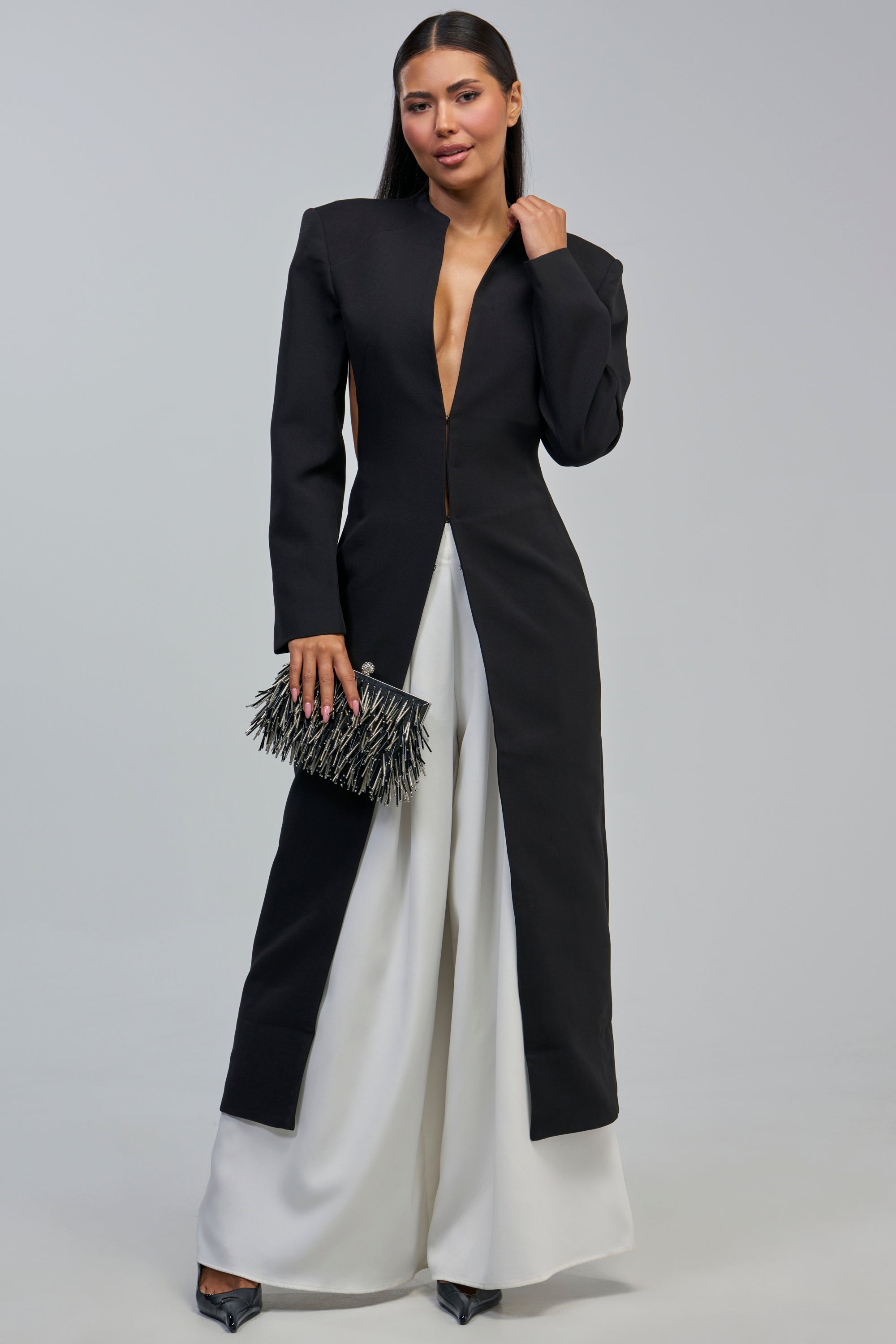 A woman stands wearing the WHEREVER YOU ARE CUTOUT TRENCH, a long black coat with a deep V neckline, styled over wide-legged white pants. She holds a black fringe clutch and wears black pointed-toe shoes against a plain, light-colored background.