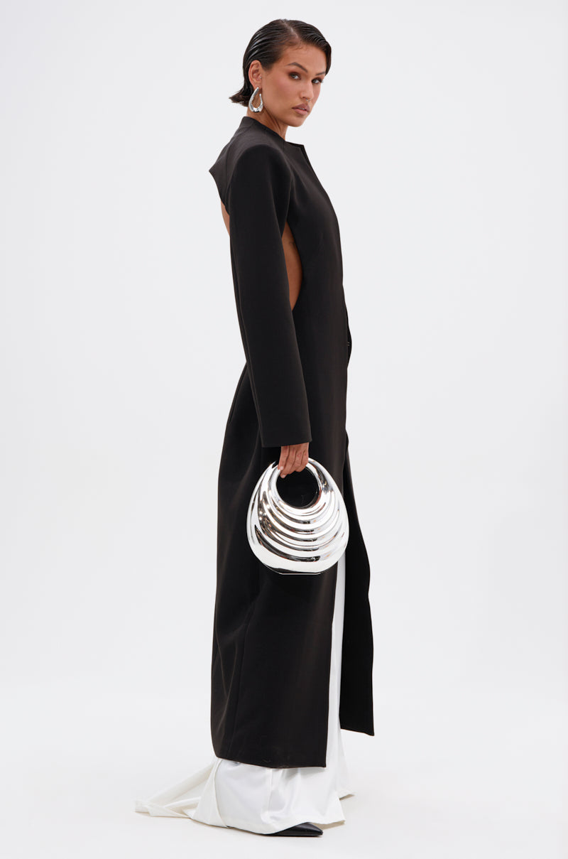 A person wearing the WHEREVER YOU ARE CUTOUT TRENCH, a long black cutout trench coat, with white trousers and a shiny sculptural silver handbag, stands against a plain white background.