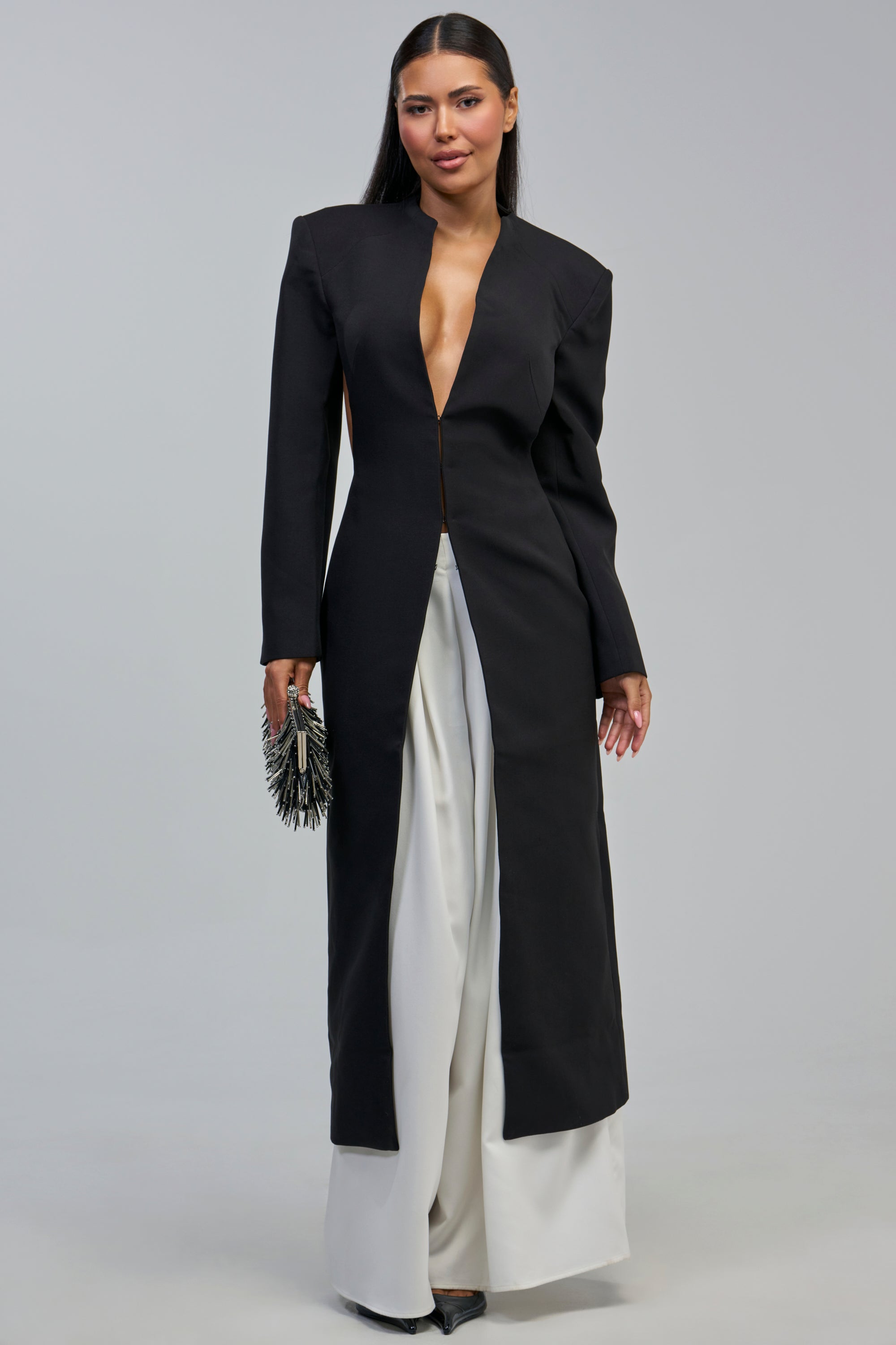 A woman stands facing forward, wearing the WHEREVER YOU ARE CUTOUT TRENCH—a long black evening coat featuring a deep neckline—paired with wide-leg white pants. She holds a spiked clutch and has long, straight hair.