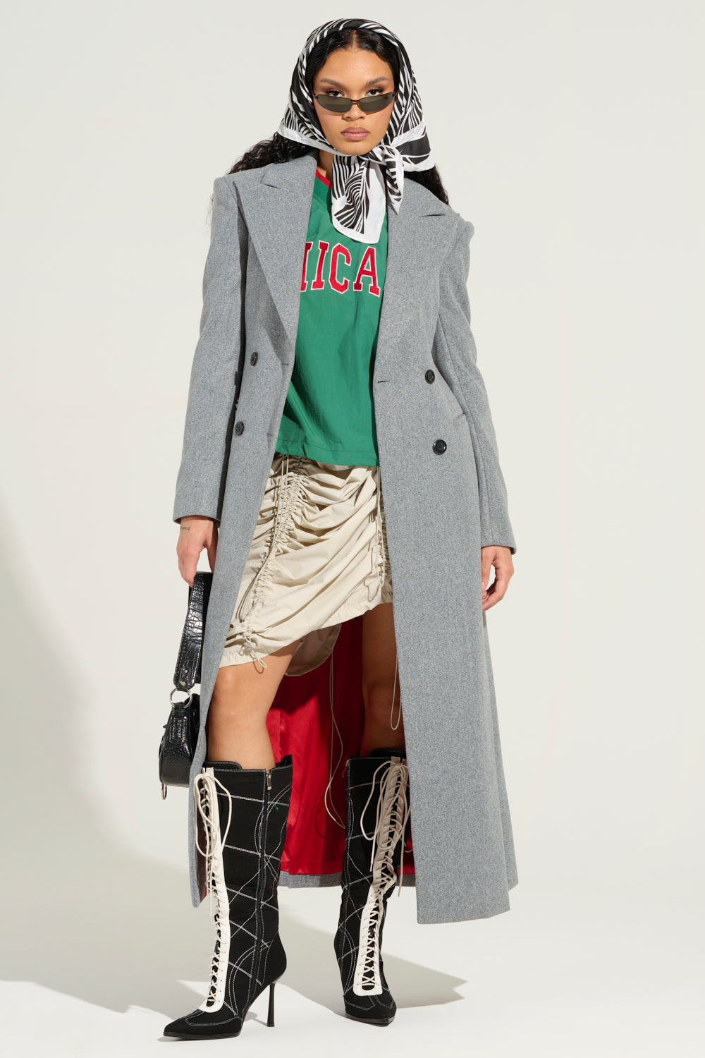 A person stylishly wears the SCARLETT LUXE TRENCH COAT, featuring a maxi longline silhouette in gray, paired with a green T-shirt adorned with red letters, a beige ruched skirt, black and white lace-up boots, and sunglasses. A zebra-patterned scarf wraps around their head as they hold a small black handbag.