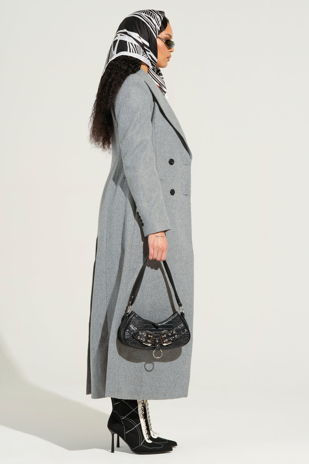 A person wearing the SCARLETT LUXE TRENCH COAT, characterized by its gray color and maxi longline silhouette, along with a black and white headscarf, sunglasses, and black boots with white laces stands in profile holding a black handbag against a plain background.
