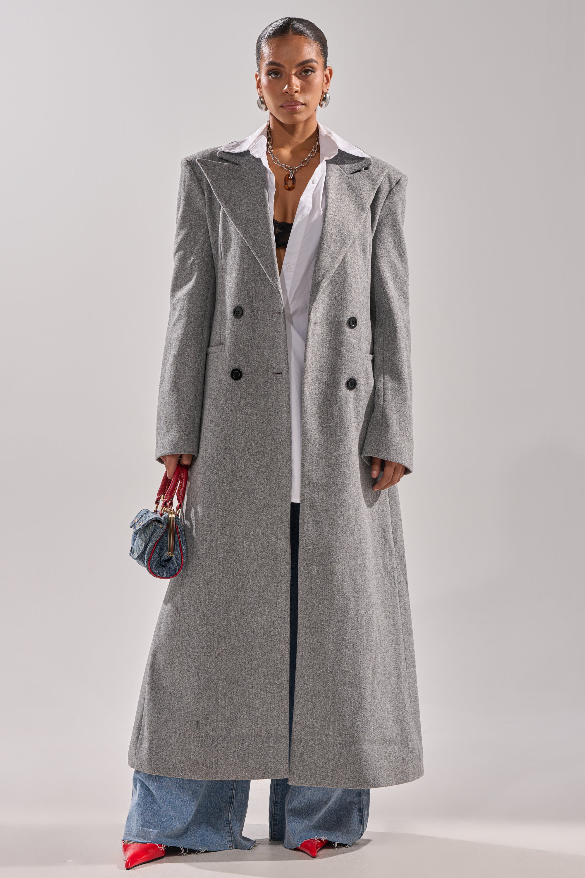 A person wears the SCARLETT LUXE TRENCH COAT over a white shirt and blue wide-leg jeans, styled with red pointed shoes, layered jewelry, and a small purse against a plain, light background. 2