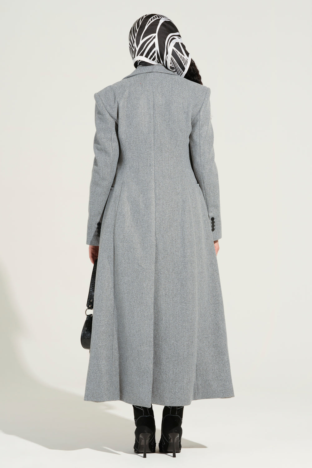 Dressed in the SCARLETT LUXE TRENCH COAT, which is double-breasted and gray, an individual stands with their back to the camera, wearing a patterned black-and-white headscarf. They hold a black handbag and wear black boots against a plain white background.