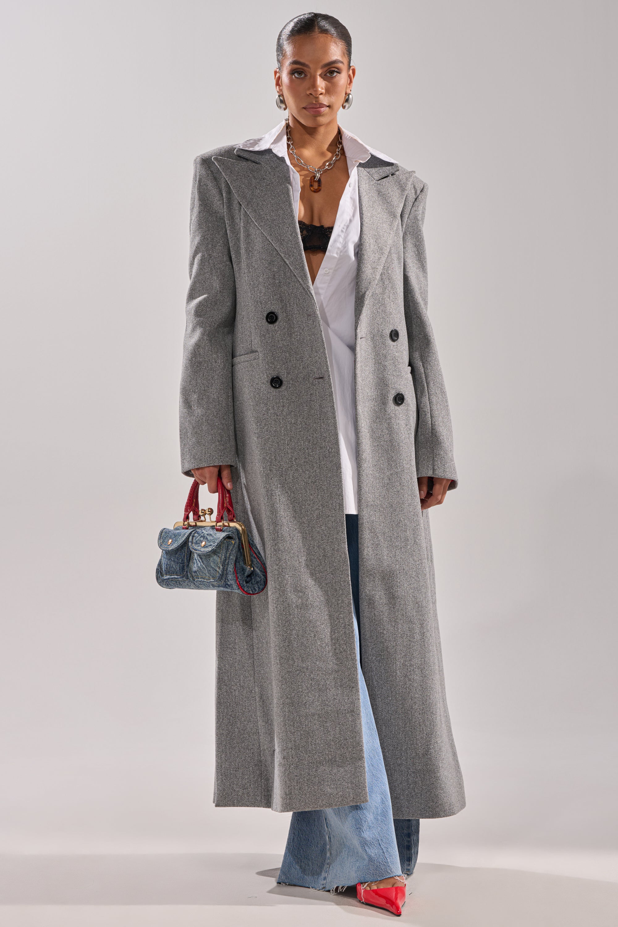 A woman wearing the SCARLETT LUXE TRENCH COAT over a white shirt and blue jeans, holding a small denim handbag with red handles, paired with red pointed-toe shoes and statement earrings. 3