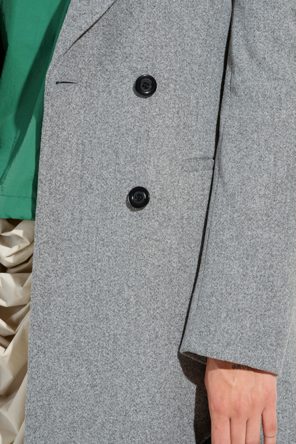 Close-up of a person wearing the SCARLETT LUXE TRENCH COAT in gray, featuring black double-breasted buttons over a green top. The trench coat elegantly settles against a creamy, slightly glossy fabric background. One hand is visible resting on the side of the coat.
