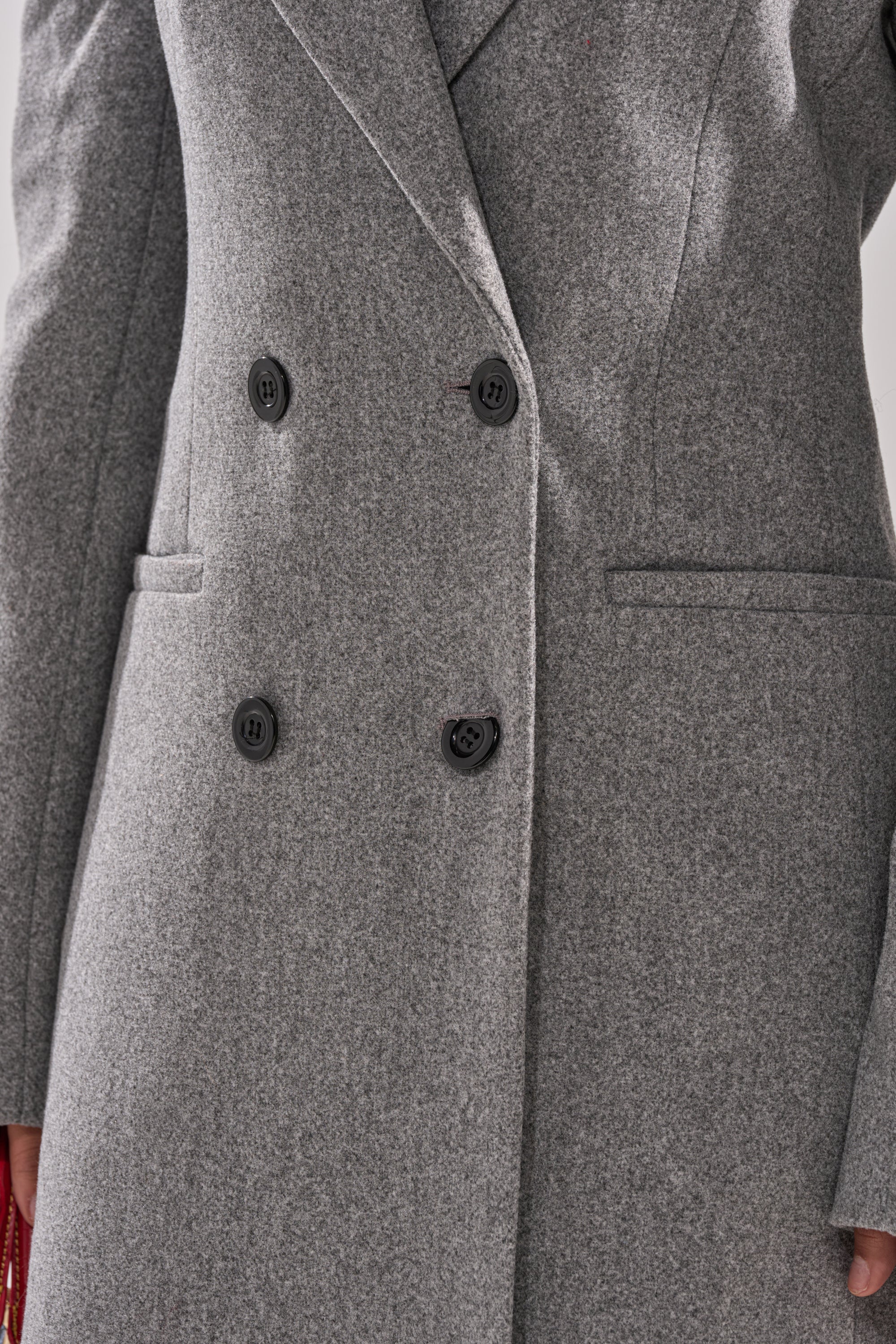 A close-up of the SCARLETT LUXE TRENCH COAT—a gray double-breasted coat with black buttons and two front pockets—worn by a person. Only the torso and part of one arm are shown. 8