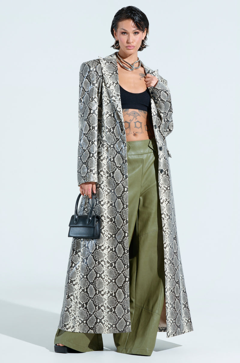 Wearing the INCOGNITO Snake Print Trench in Ivory with a longline silhouette, a black crop top, olive green wide-leg pants, and holding a small black handbag, the person stands confidently against a plain light-colored background.