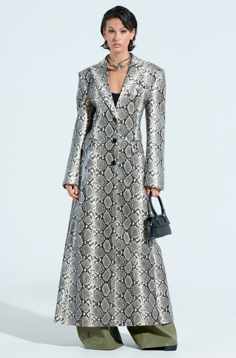 A person stands confidently against a plain background, dressed in the INCOGNITO Snake Print Trench in Ivory and wide-legged green pants. They hold a small black handbag, have short dark hair, and exude a serious expression.
