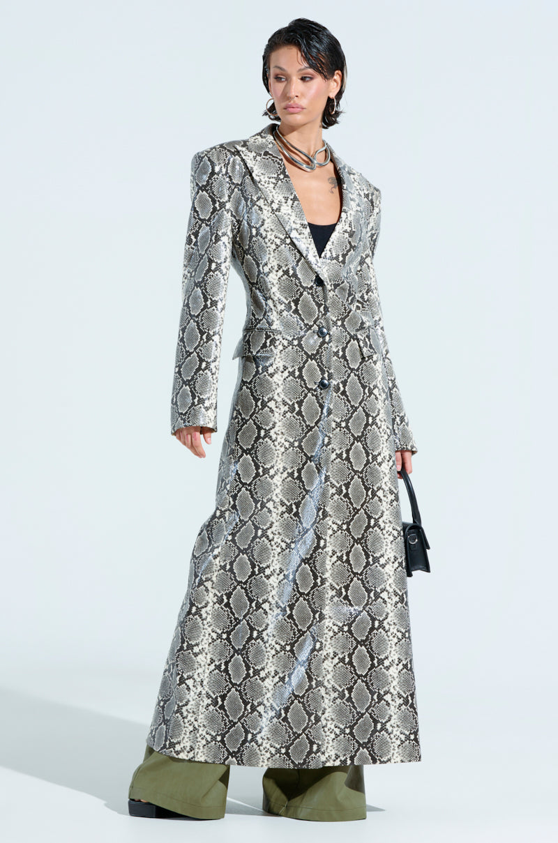A person in an INCOGNITO Snake Print Trench in ivory and olive green platform boots stands confidently. A small black handbag and layered necklaces accentuate their short dark hair, with a plain, light-colored background highlighting their striking longline silhouette.