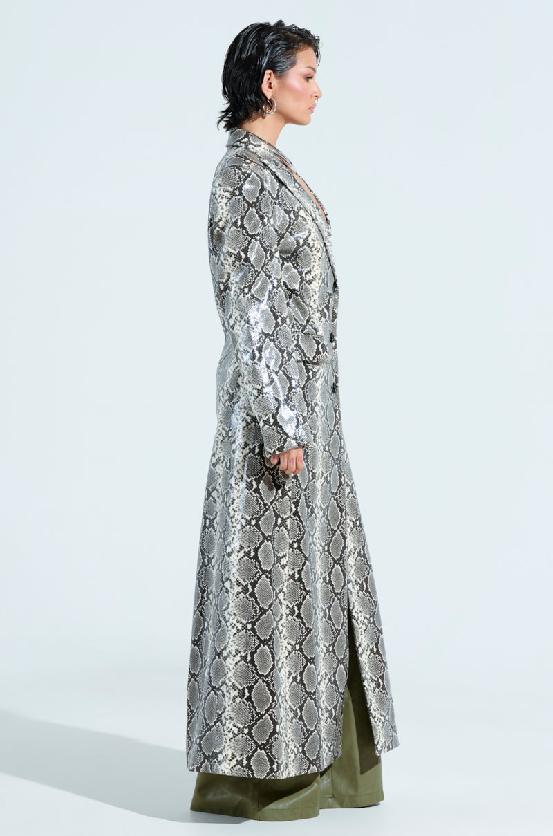 A person stands in profile wearing an INCOGNITO SNAKE PRINT TRENCH IN IVORY with a longline silhouette, paired with olive green pants. The light-colored background enhances the outfit's striking details.