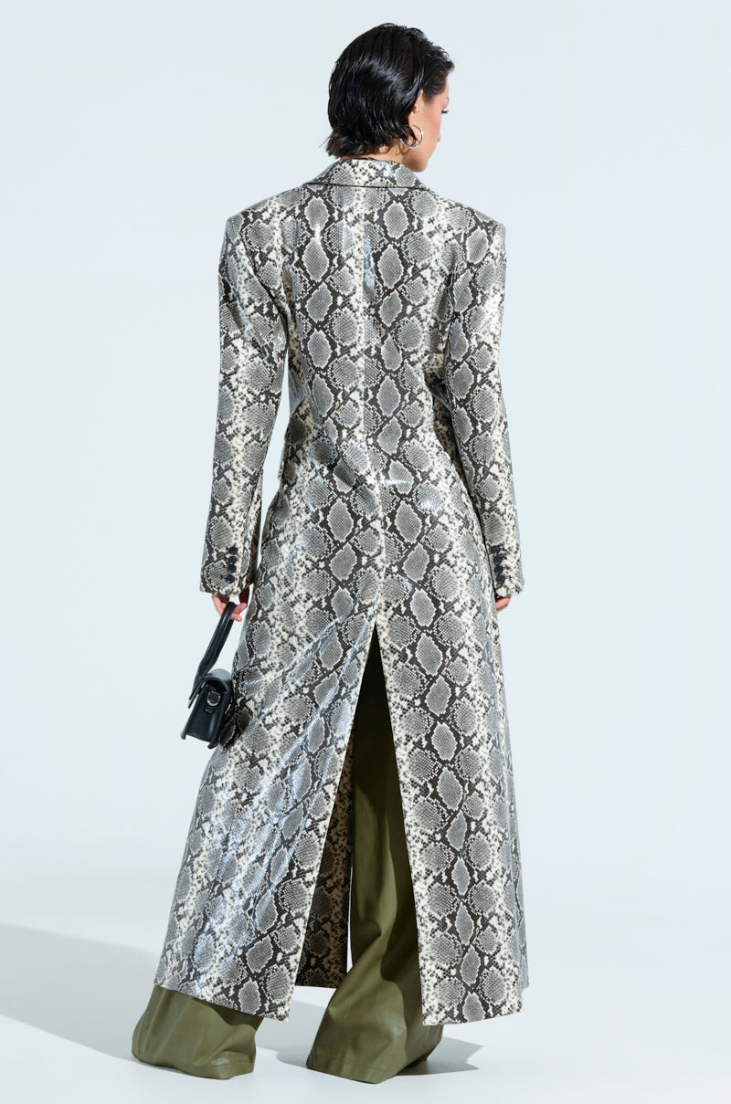 A person with short, dark hair is wearing an ivory snake print trench coat with a longline silhouette and high slit in the back, over olive green pants. They are holding a small black purse, standing against a light background.