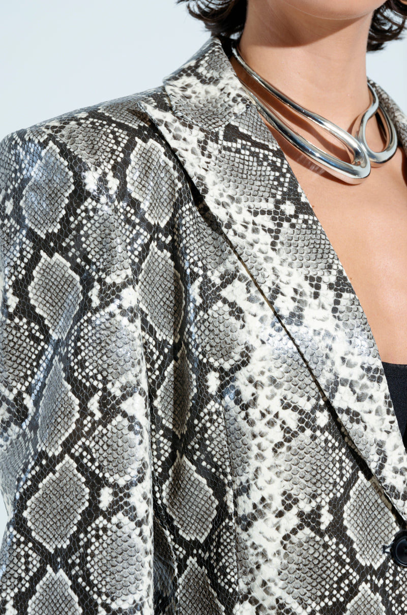 A person wearing the Incognito Snake Print Trench in Ivory paired with a silver chunky necklace exemplifies contemporary style with short, dark hair. The longline silhouette enhances sophistication against a plain white or light gray background.