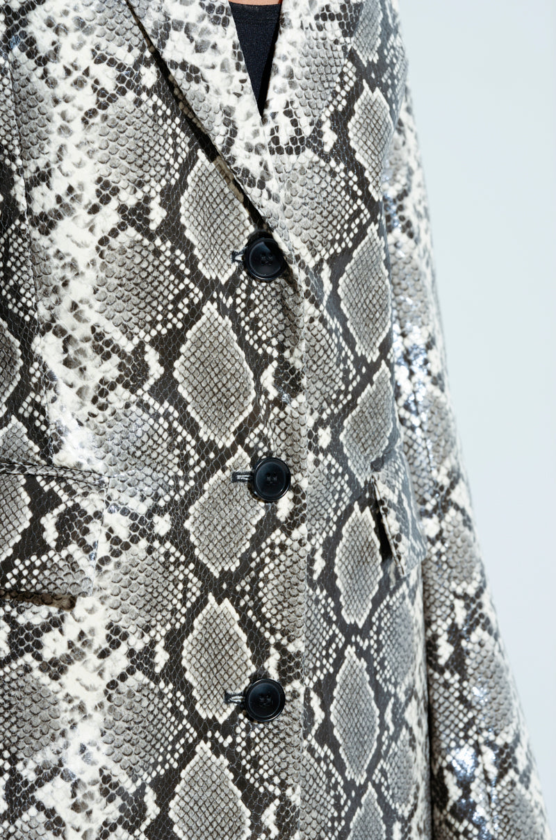 The INCOGNITO SNAKE PRINT TRENCH IN IVORY features a snakeskin pattern with black buttons. Its fabric displays intricate gray, white, and black scales, delivering a textured and stylish look reminiscent of a longline silhouette.
