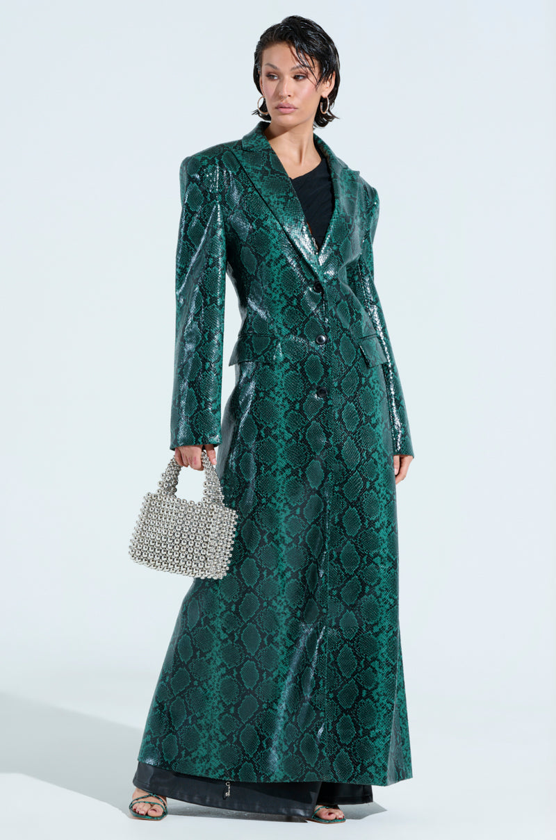 A person poses against a plain white background wearing the INCOGNITO SNAKE PRINT TRENCH IN GREEN over a black outfit, complemented by a small beaded handbag. Their hair is styled short and wet.