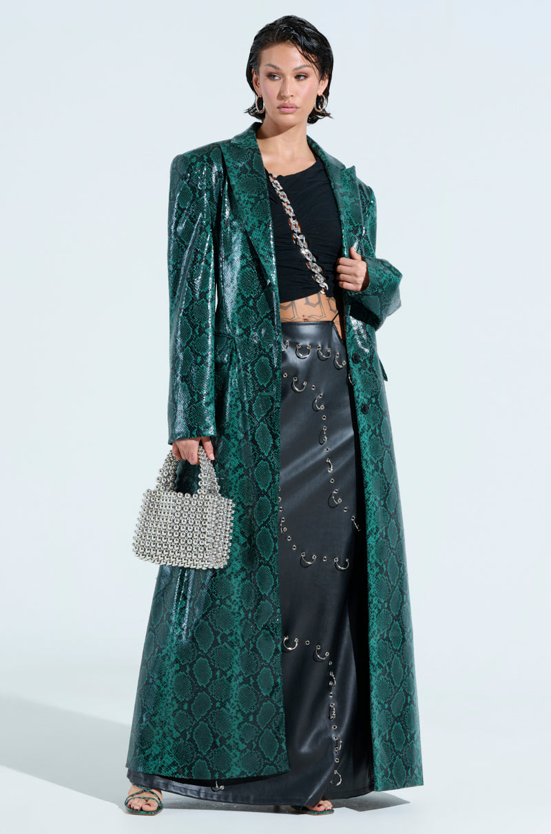A person poses in the INCOGNITO SNAKE PRINT TRENCH IN GREEN over a black crop top and metal-accented black skirt, holding a silver beaded handbag and wearing sandals against a plain backdrop.