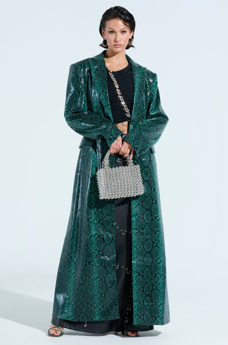 A person is wearing the INCOGNITO SNAKE PRINT TRENCH IN GREEN over a black outfit. They're holding a beaded handbag with wet-look hair against a plain white background.