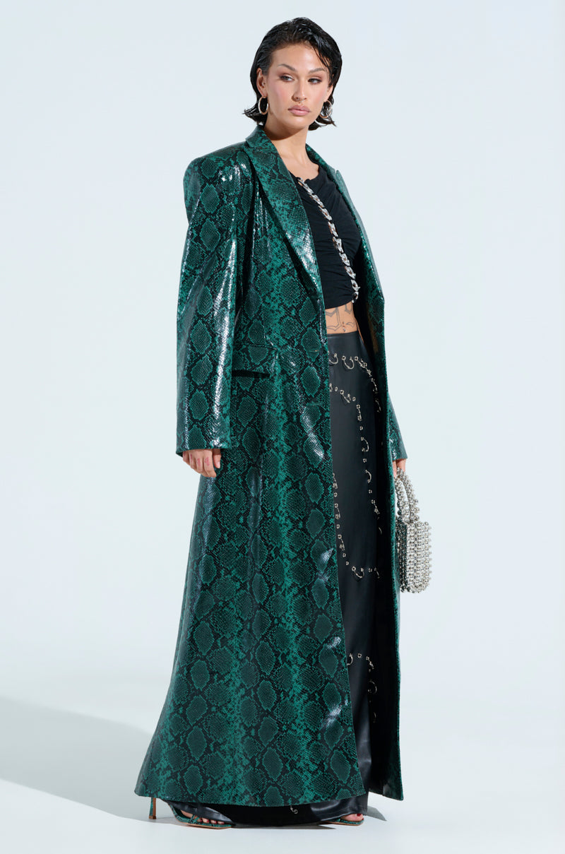 A person in an INCOGNITO SNAKE PRINT TRENCH IN GREEN over a black outfit stands against a plain backdrop, holding a small beaded handbag and wearing black heeled sandals. Their sleek, wet-styled hair perfectly complements the bold trench choice.