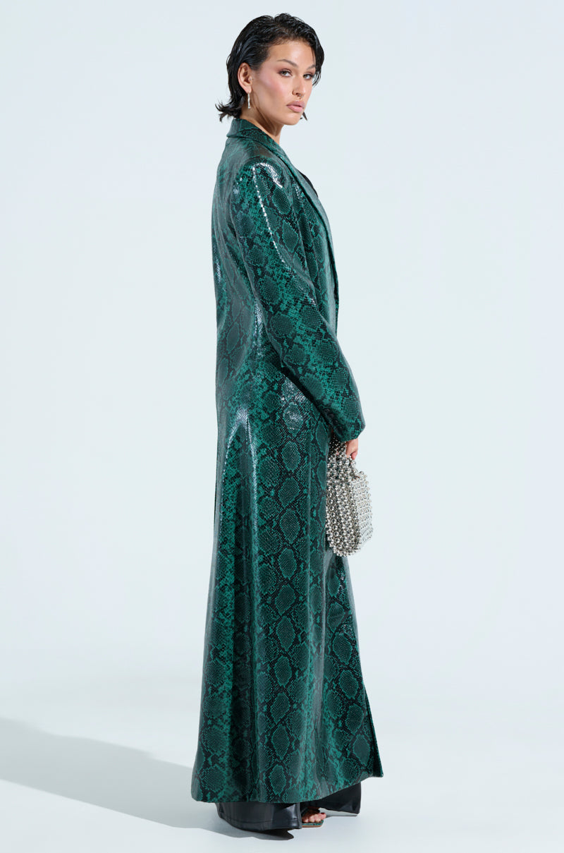 Wearing the INCOGNITO SNAKE PRINT TRENCH IN GREEN, a person poses against a plain background, holding a small studded handbag and sporting short, dark hair.