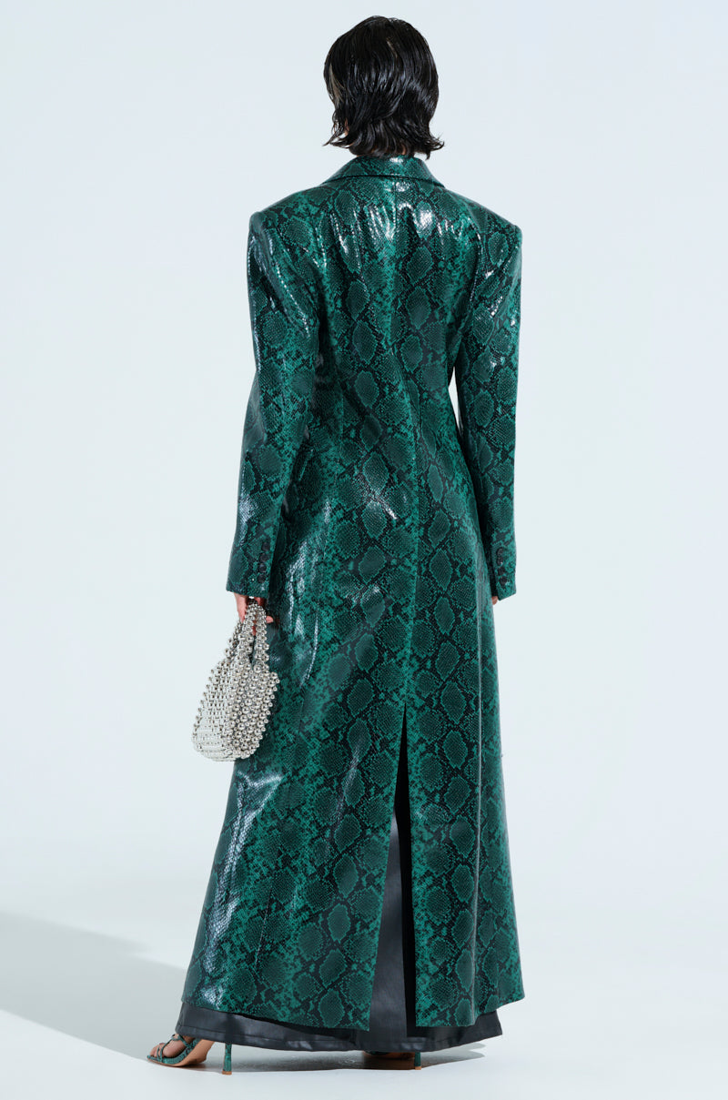 Facing away from the camera, a person wears an INCOGNITO SNAKE PRINT TRENCH IN GREEN, featuring broad shoulders. They hold a small beaded handbag and wear sandals, all on a pristine white backdrop.
