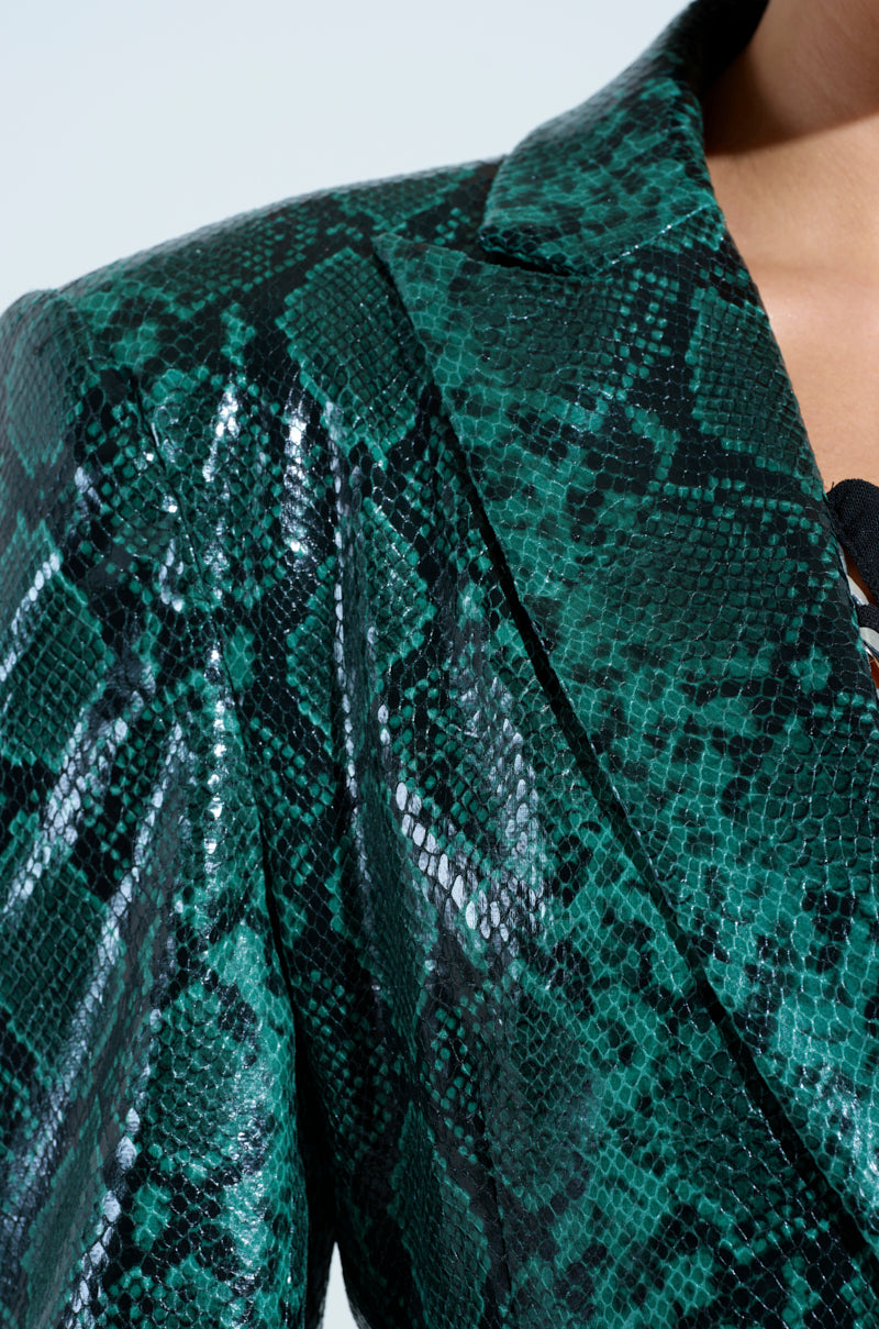 Close-up of a person wearing the INCOGNITO SNAKE PRINT TRENCH IN GREEN. The shiny faux leather jacket showcases a vivid reptile-like design and sharp lapel, capturing part of the person’s neck and shoulder in the image.