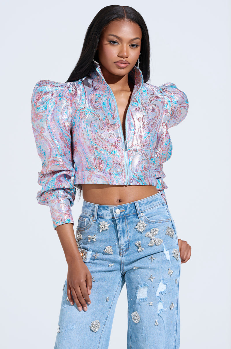 A woman wears the colorful DELILAH PUFF SHOULDER BOMBER with a shiny puff shoulder and paisley pattern, paired with light blue floral embellished jeans. The simple background enhances her striking metallic style.