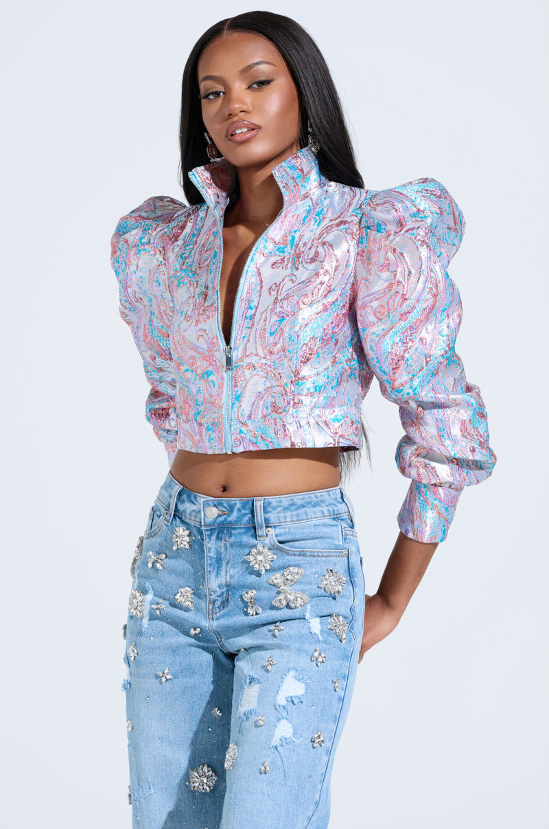 A person boldly wears the DELILAH PUFF SHOULDER BOMBER, a vibrant brocade-patterned jacket with puff sleeves, against a simple backdrop. Pairing it with light blue jeans adorned in metallic florals and sleek, long hair completes their striking look.